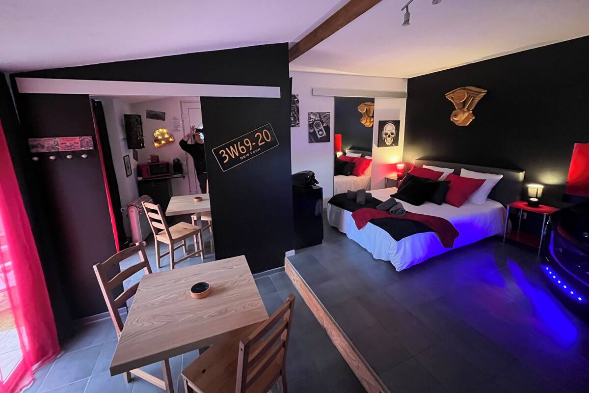 A compact hotel room with a bed, a desk, and a dining area. There is a sign on the wall indicating the room number and location.