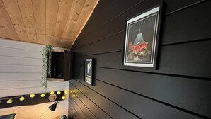 Une pièce aux murs et au plafond en bois foncé, avec un petit coin salon comprenant un banc et une table. Des tableaux encadrés ornent les murs et une guirlande lumineuse est disposée sur le sol.