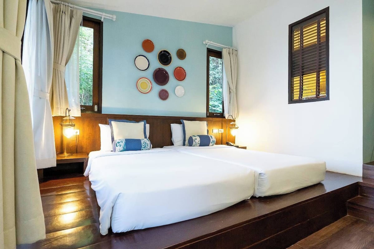 A double bed with white linens, two bedside tables with lamps, a wall-mounted clock, and a window with blinds.