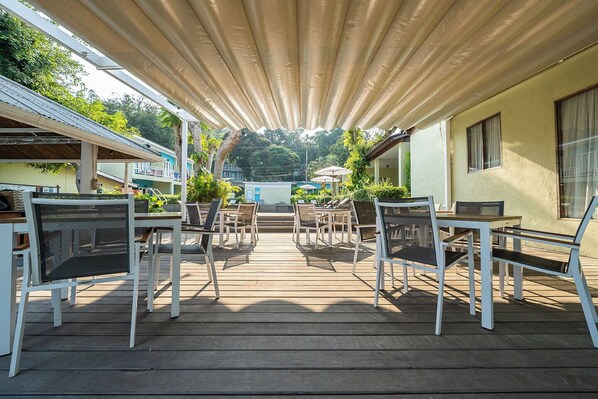 Un patio al aire libre con mesas y sillas de madera, un área cubierta con toldo y vistas a un edificio y árboles al fondo.
