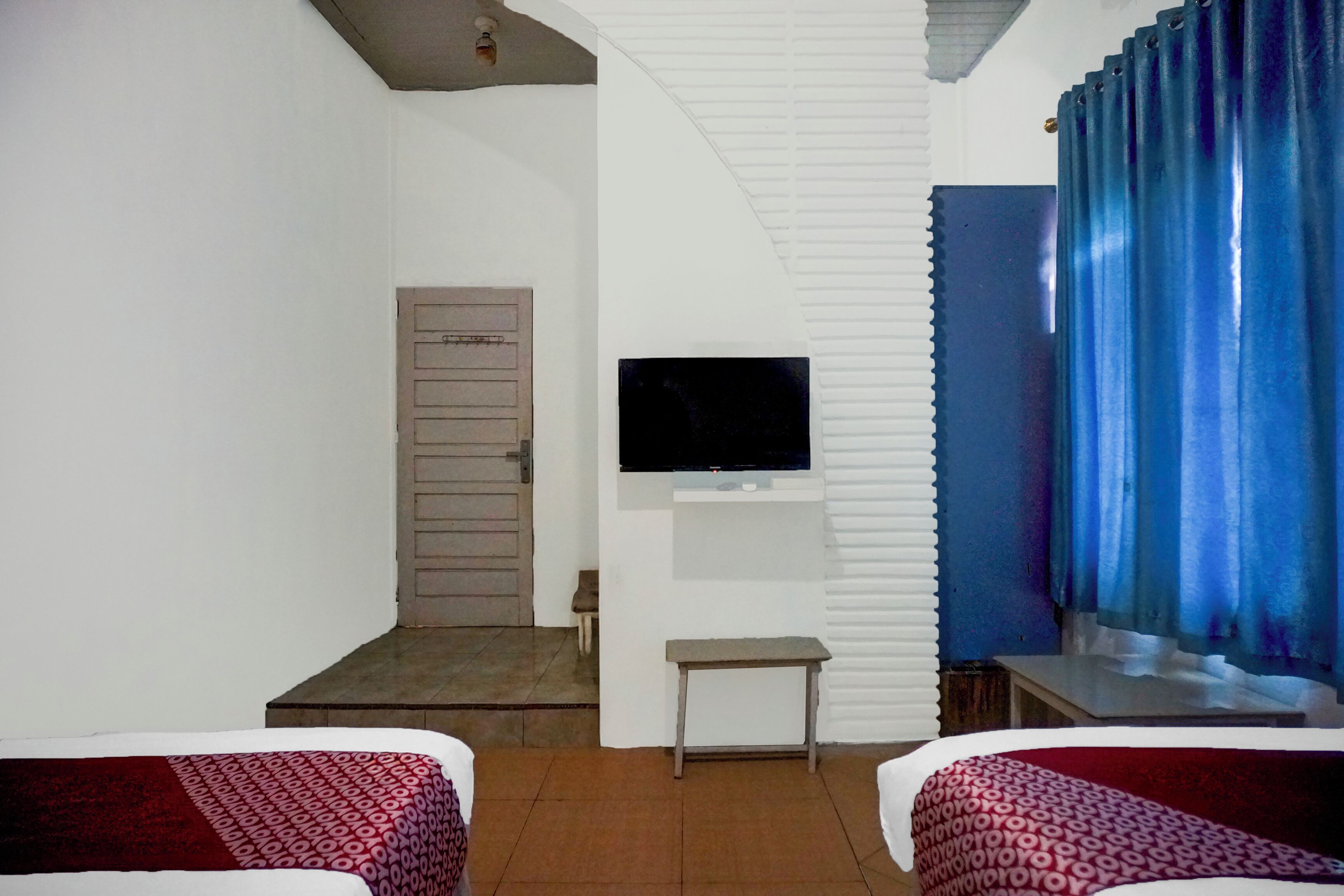 Ein Hotelzimmer mit zwei Betten, einem an der Wand befestigten Fernseher, einem kleinen Tisch und einer Tür zu einem weiteren Zimmer.