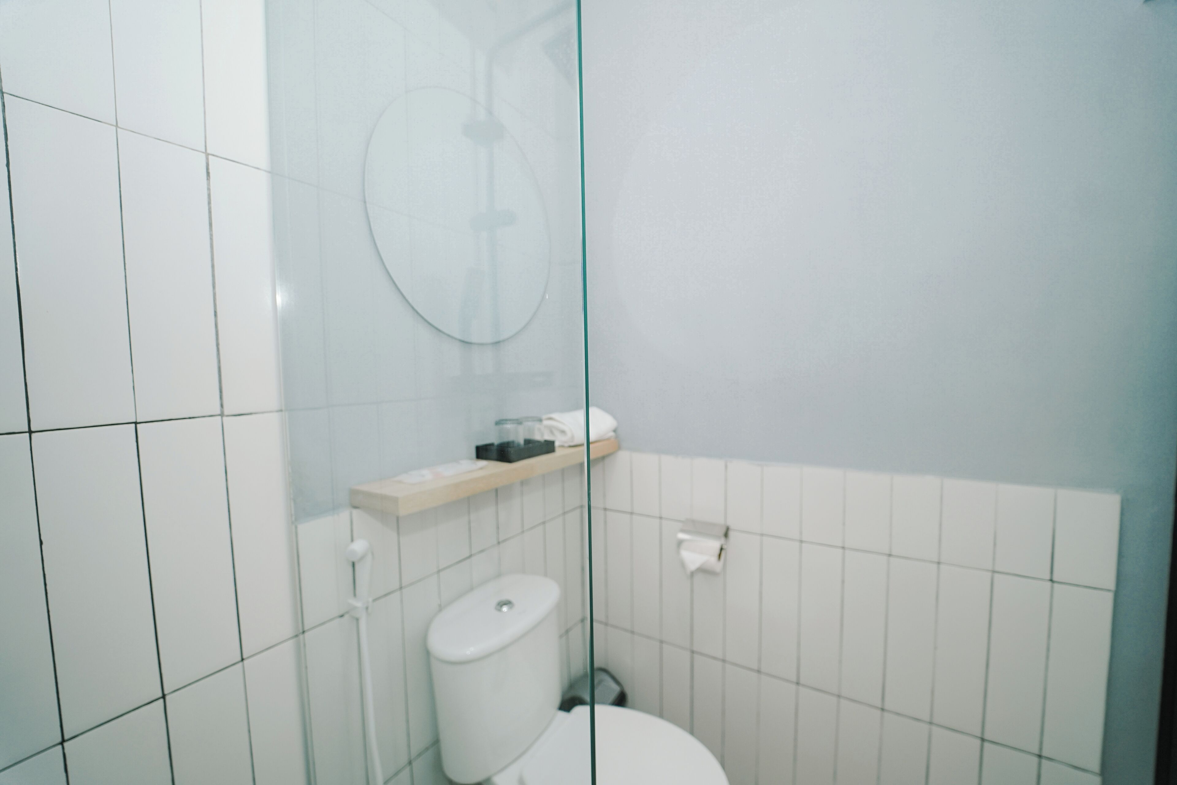 Un baño pequeño y moderno con baldosas blancas, un inodoro, cabina de ducha con mampara de cristal y un estante con artículos de aseo.