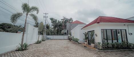 A paved courtyard with a white building featuring a red roof, surrounded by plants and a white wall.