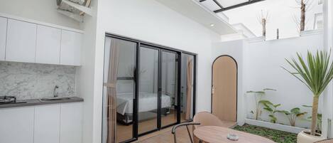 Habitación de hotel compacta con kitchenette, zona de comedor con mesa y sillas, un amplio ventanal corredero que da a un balcón y una planta en maceta.