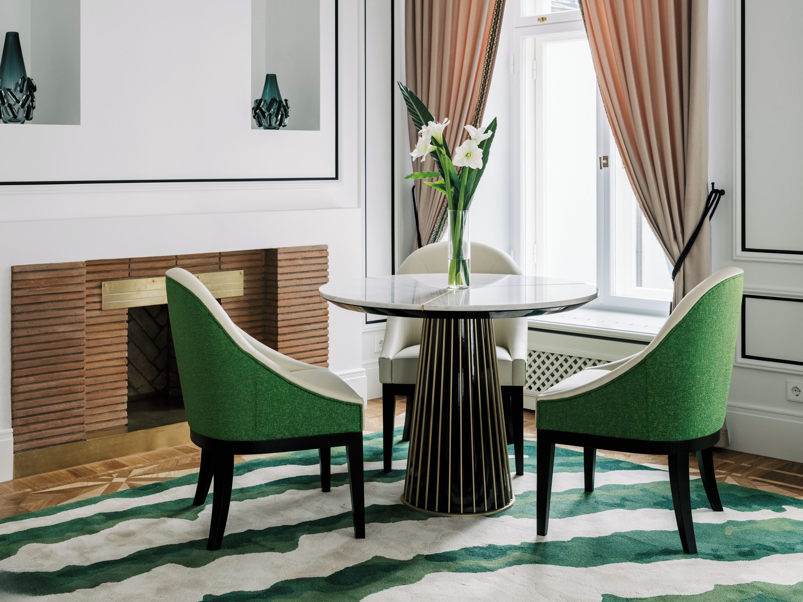 A dining area with a round table, two green chairs, a vase with flowers, and a fireplace.