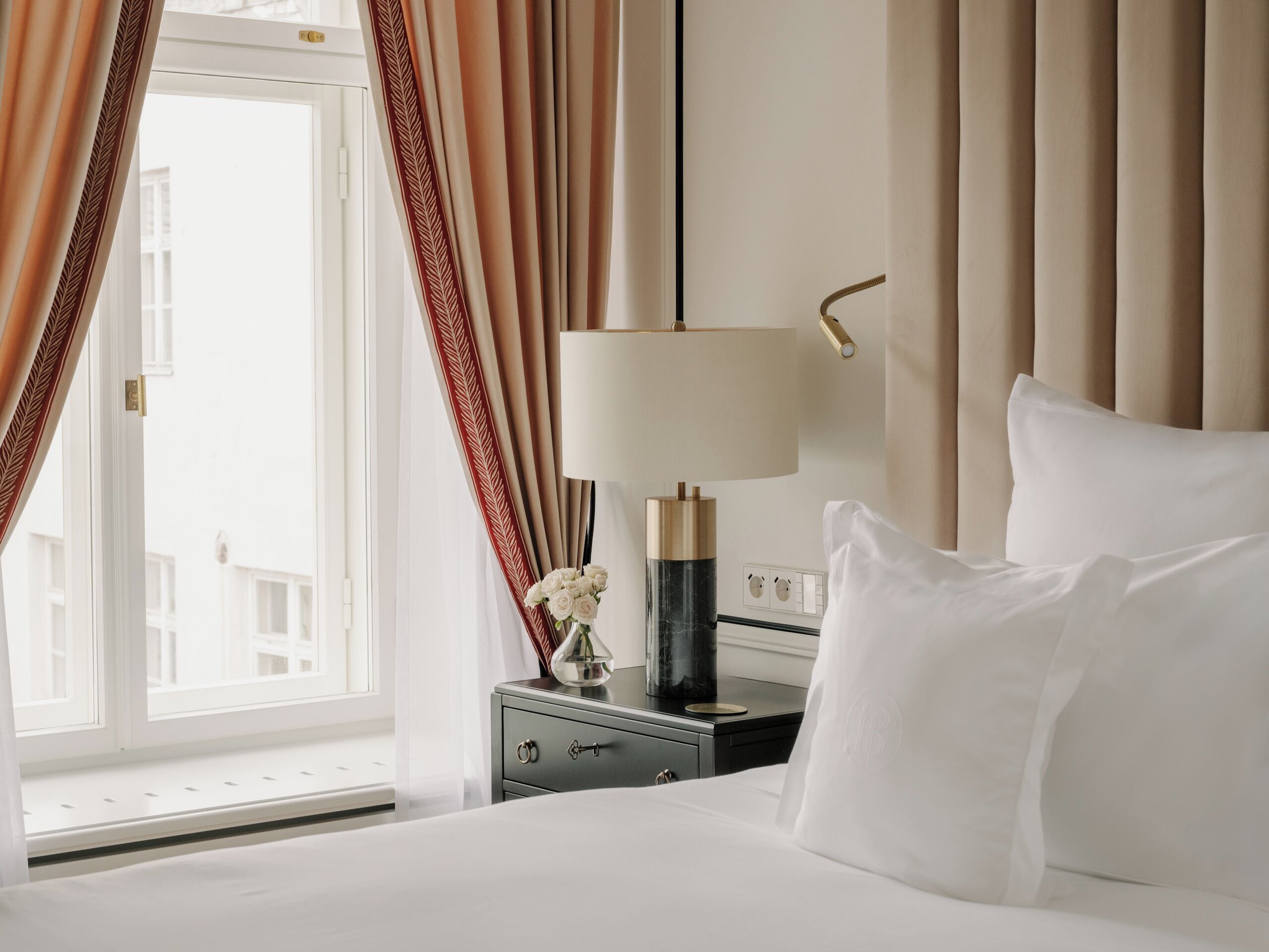 Quarto de hotel com cama, mesa de cabeceira com luminária, um vaso com flores e vista para prédios.