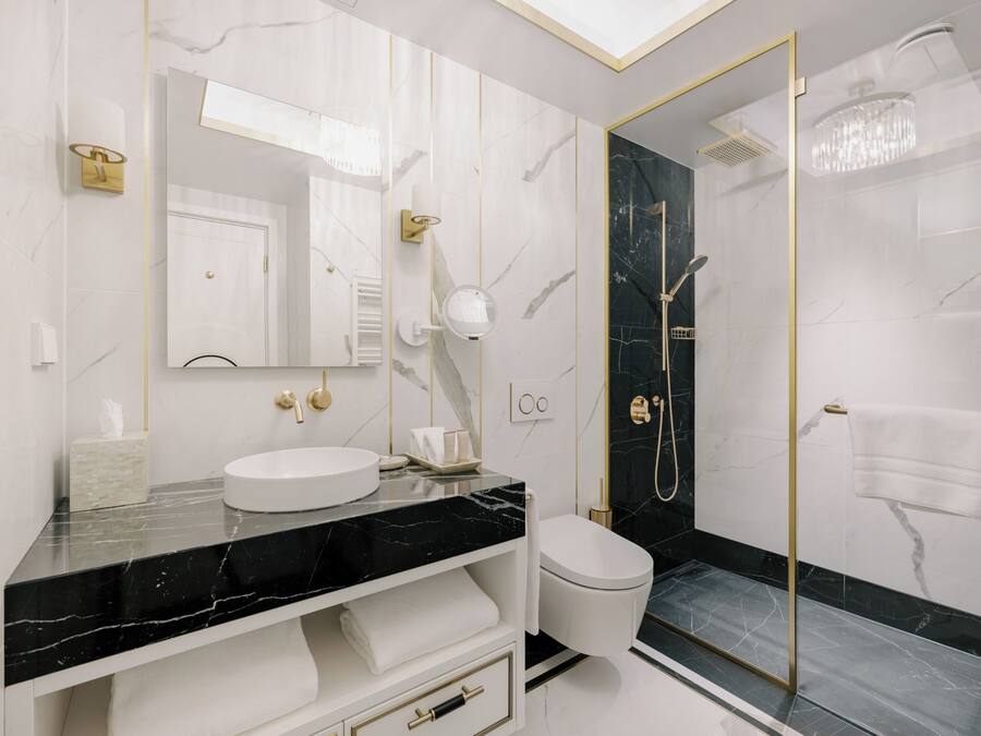 Un baño moderno con tocador de mármol, lavamanos blanco, inodoro y ducha con mampara de vidrio y marco dorado.