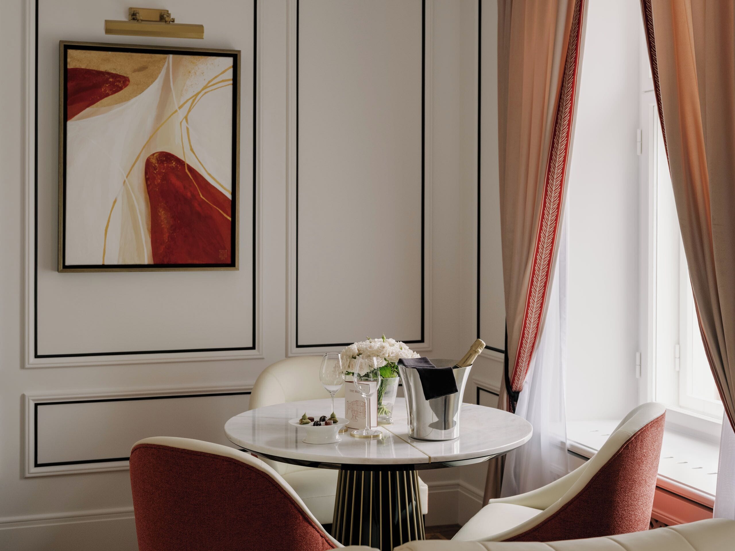 Un coin repas avec une table ronde, des chaises blanches, un tableau abstrait encadré et une fenêtre avec des rideaux.