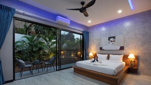 Une chambre moderne avec un grand lit, deux tables de chevet, un ventilateur de plafond et une vue sur un espace extérieur aménagé avec des chaises et des plantes, accessible par une porte-fenêtre coulissante.