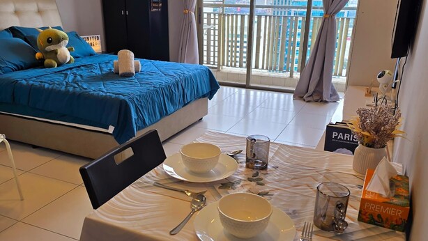 A hotel room with a bed, a dining table set with plates and bowls, a chair, and a view of a cityscape through large windows.