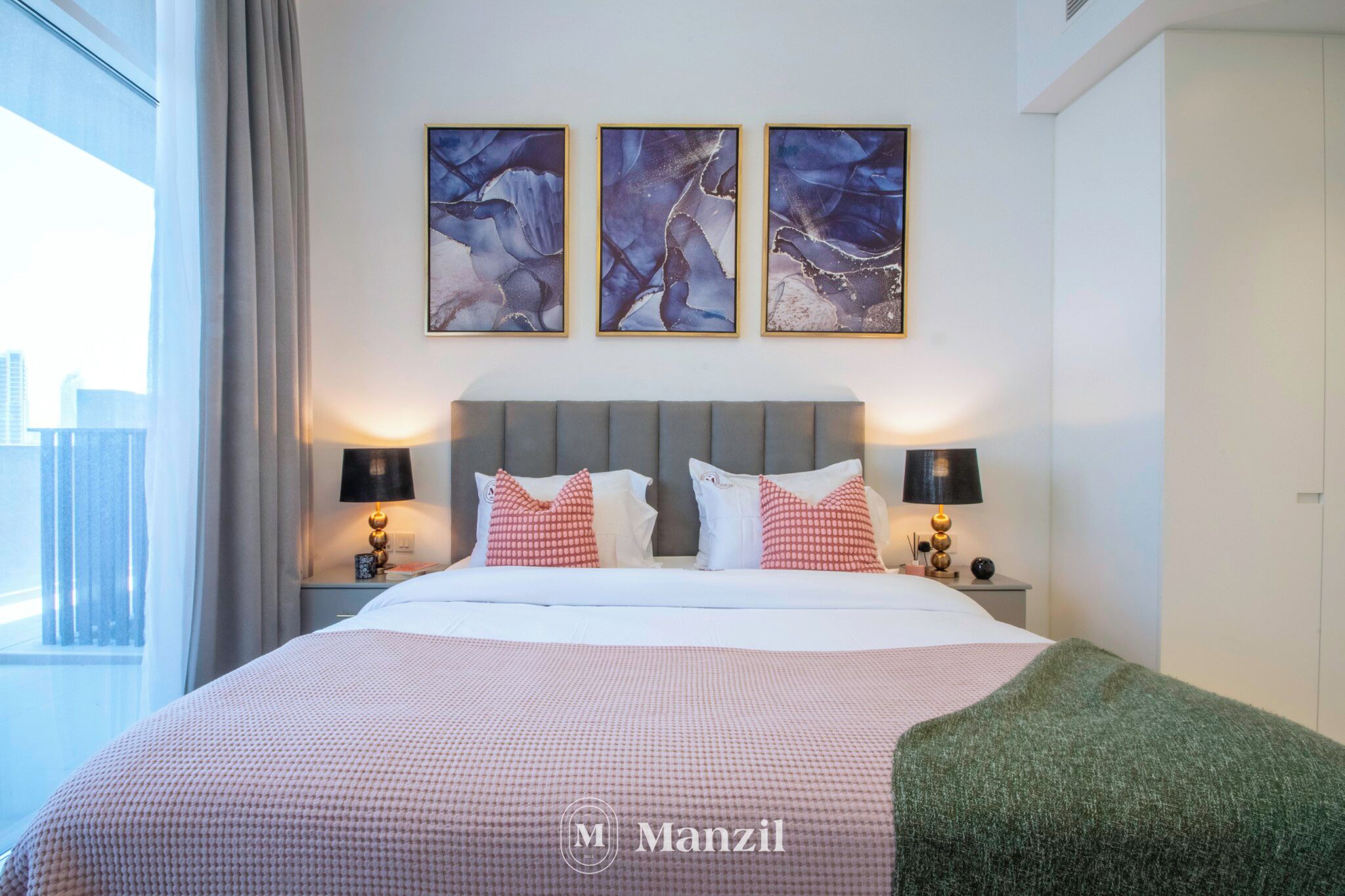 A hotel room with a large bed, two bedside tables with lamps, a grey headboard, and three abstract paintings on the wall.