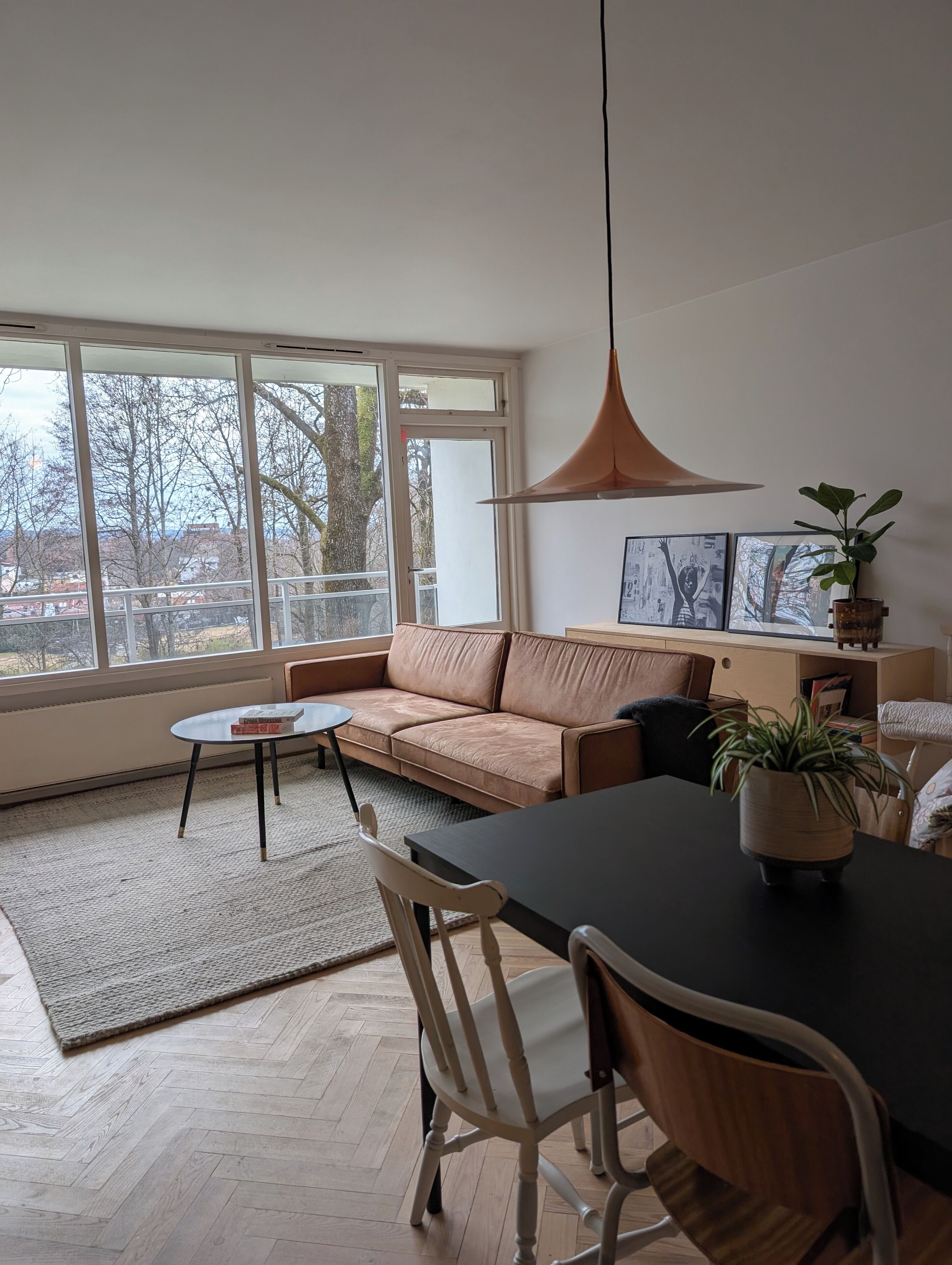 Een moderne woonkamer met een bank, eettafel, stoelen, een hanglamp en uitzicht door grote ramen.
