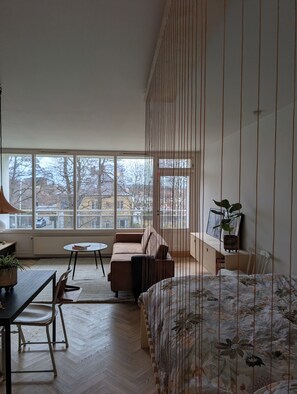 A modern living room with a sofa, a dining table, a small round table, and a bed with floral bedding. There are large windows providing a view of the outside.