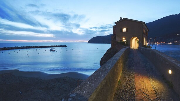 Un bâtiment situé sur la côte, avec un chemin de pierre menant à son entrée, offrant une vue sur une mer calme et des montagnes lointaines.
