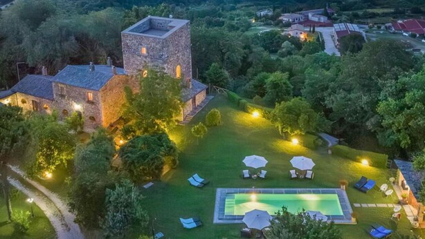 Un edificio de piedra con una torre, rodeado de un jardín bien iluminado que incluye piscina, sillas de lounge y sombrillas.