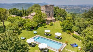 Zona de piscina con sillones de descanso y sombrillas, rodeada de exuberante vegetación y un edificio de piedra con torre.