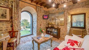 Una habitación rústica con paredes de piedra, techo de madera, candelabro, televisor, chimenea y vistas al exterior.
