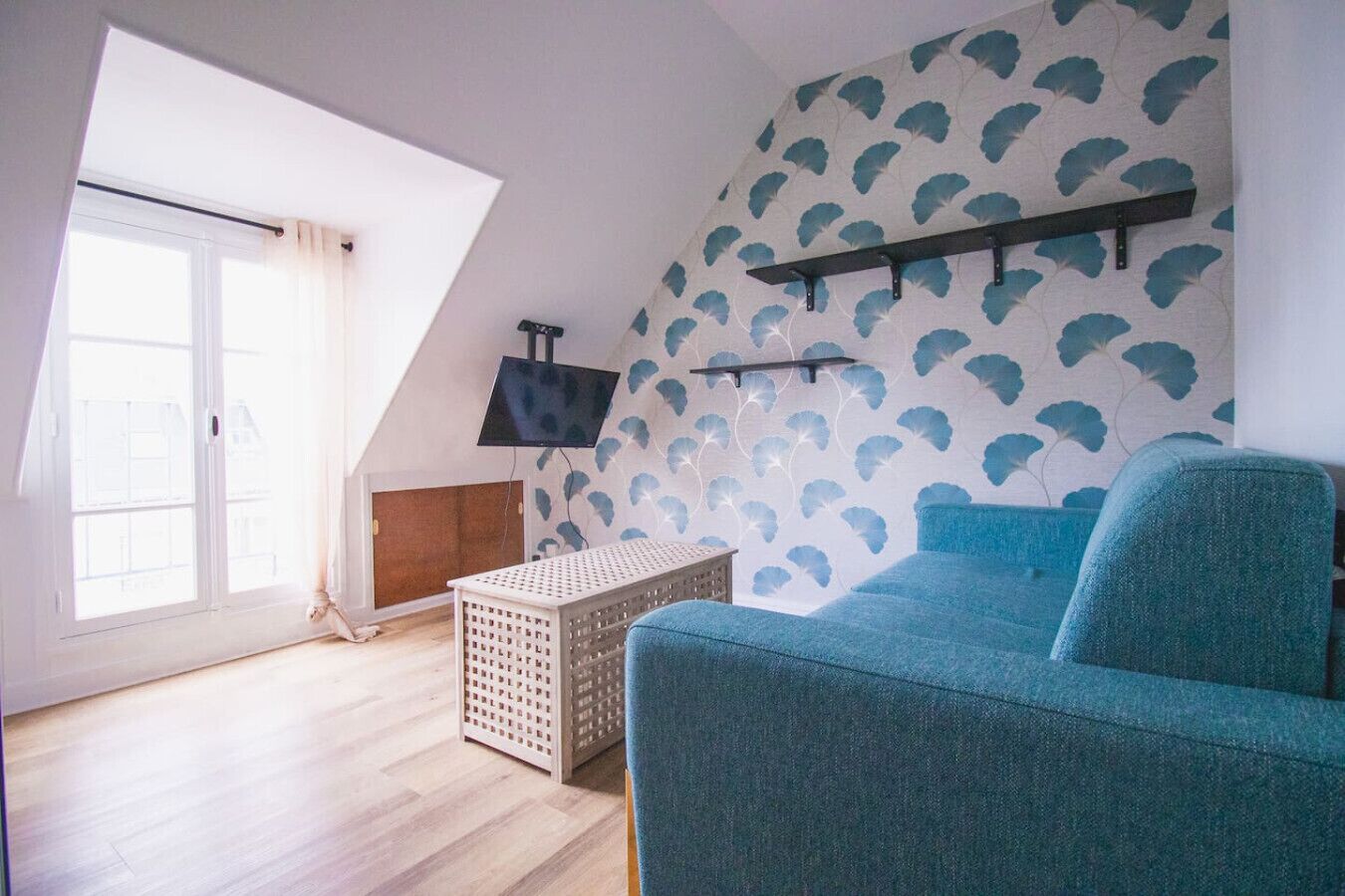 A room with a sloped ceiling, a blue sofa, a TV on the wall, and a window with curtains.
