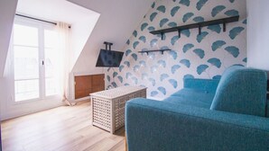 A room with a sloped ceiling, a blue sofa, a TV on the wall, and a window with curtains. - Studio FullEquiped 2person with balcony 17th (Paris)
