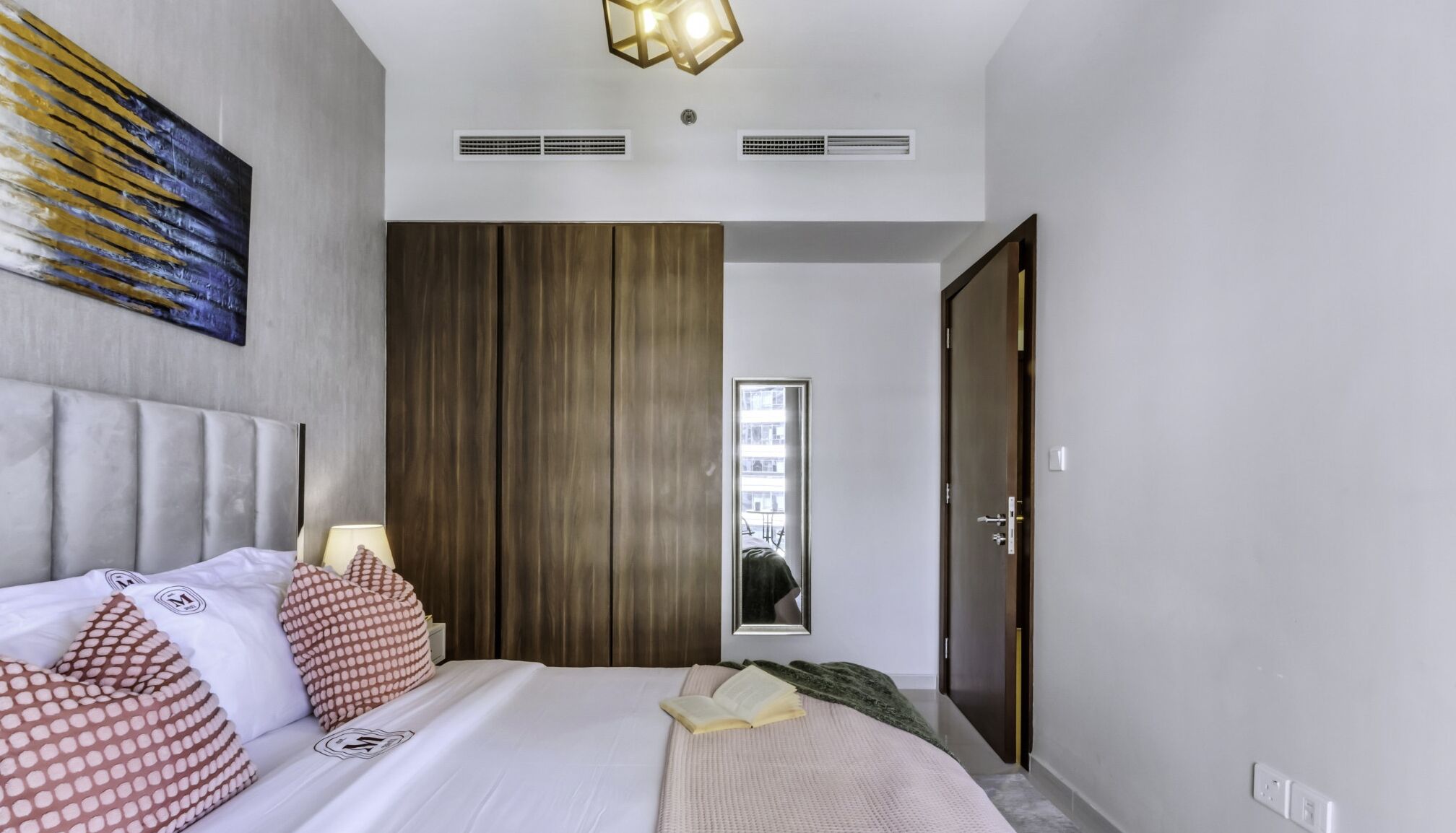 Une chambre d’hôtel moderne avec un grand lit, une tête de lit, un mur recouvert de panneaux en bois, un miroir et une porte donnant sur un balcon.