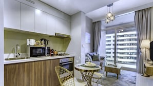 A compact kitchen with modern appliances, a dining area with a round table and chairs, and a large window offering a view of a high-rise building. - Manzil-1BR w Canal View near Dubai Mall (Dubai)