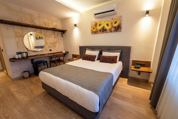 A hotel room with a large bed, a wooden headboard, a mirror, a wall-mounted air conditioner, a small table with a kettle, and a shelf with toiletries.