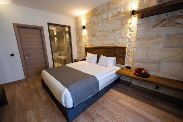 A bedroom with a large bed, wooden headboard, stone wall, and a small wooden shelf with a red flower arrangement. - Stones of Cappadocia (Nevsehir)