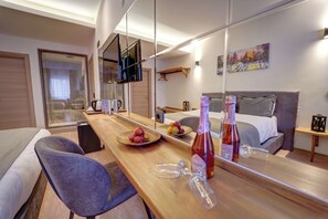 Moderni hotellihuone, jossa on puinen ruokapöytä, harmaalla päätyosalla varustettu sänky, suuri peili, joka heijastaa huonetta, ja taulu seinällä.