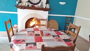 A dining area with a round table covered by a patchwork tablecloth, wooden chairs, a clock above a fireplace, and a blue and white wall. - Family hotel (Soullans)