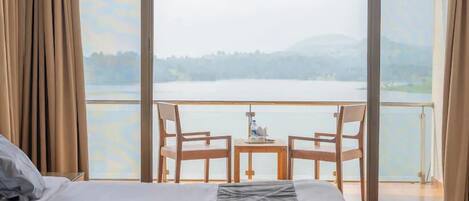 A hotel room with a large bed, two chairs, a small table, and a view of a lake through large windows.