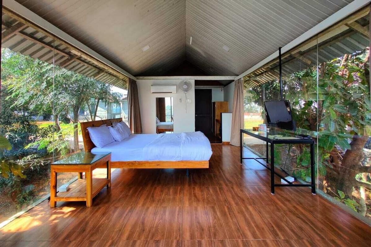 A modern bedroom with a wooden floor, a bed, a small table, and a glass wall with plants.