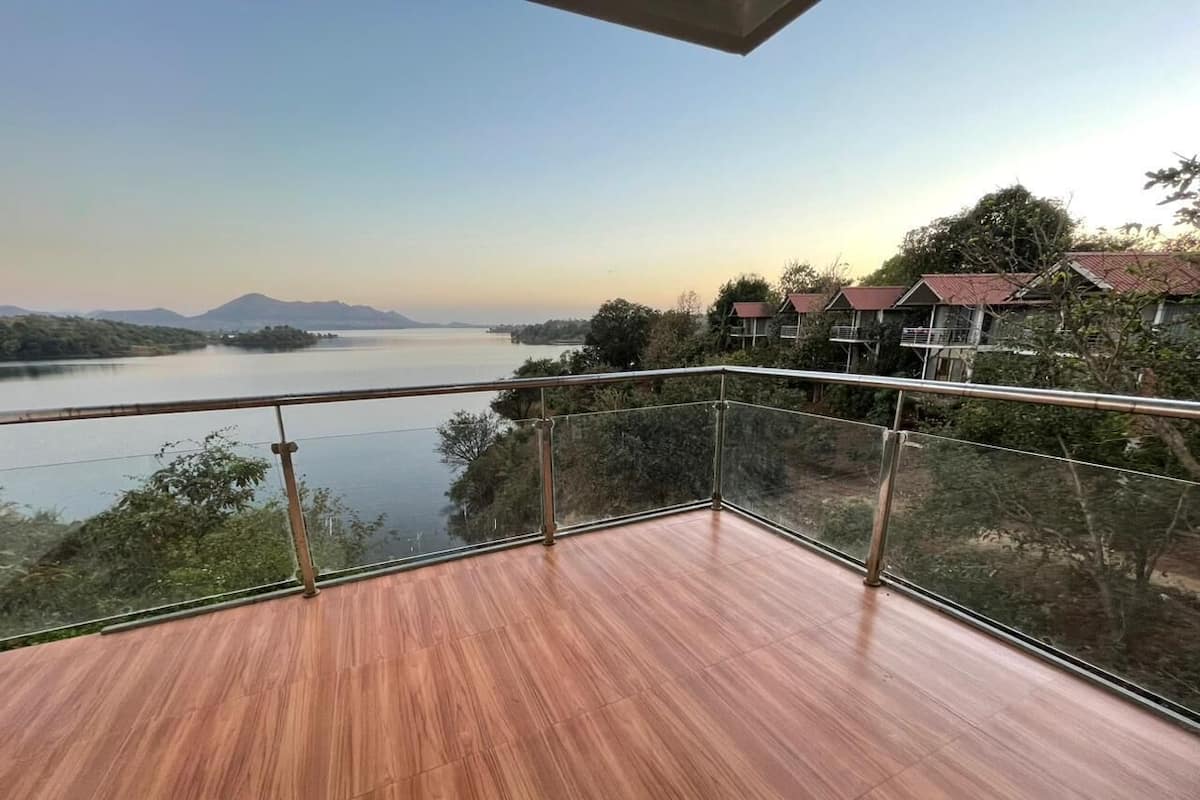 A balcony with a wooden floor and glass railing, overlooking a lake and distant mountains.