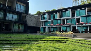 Ein modernes Gebäude mit mehreren Balkonen und großen Fenstern, umgeben von einer gepflegten Rasenfläche und einer Außenbereich-Sitzgelegenheit mit Sonnenschirmen.