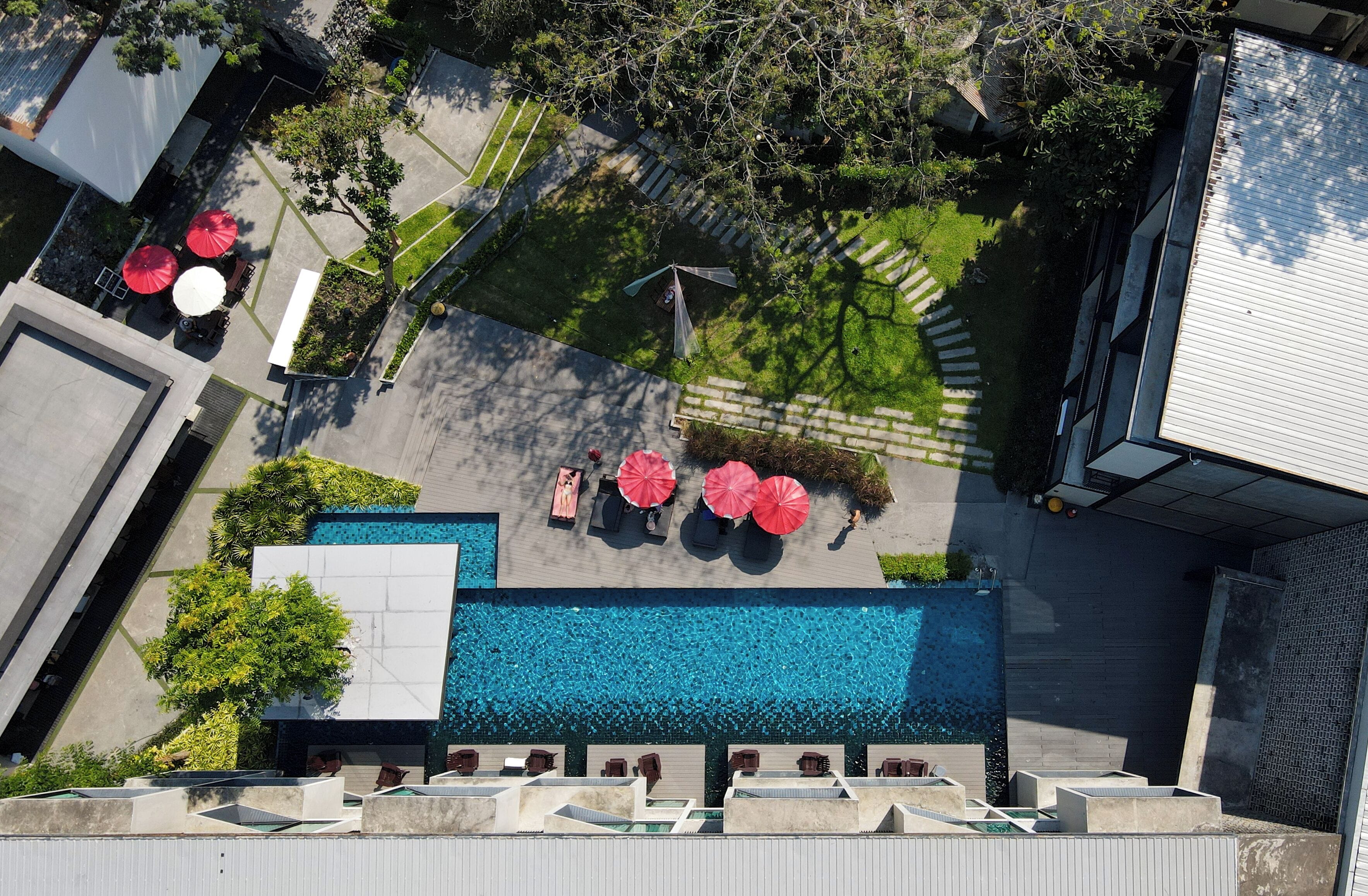 An aerial view of a rooftop pool area with sun loungers and umbrellas, surrounded by greenery and a clear blue pool.