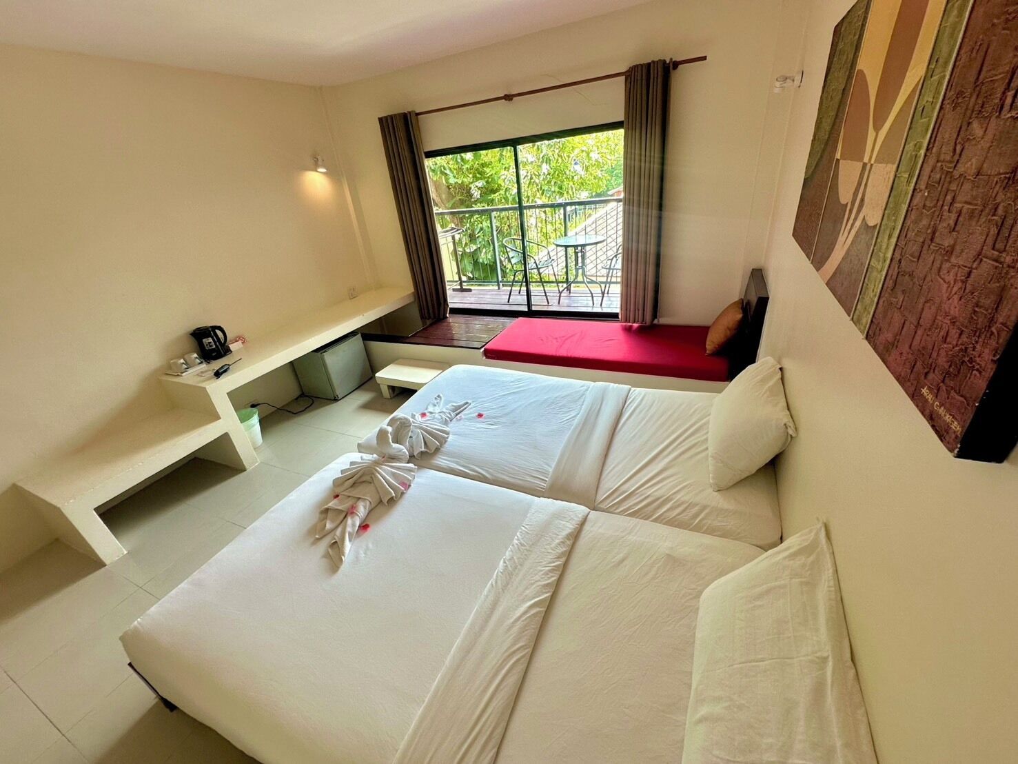 Une chambre d’hôtel avec deux lits, un petit bureau, une fenêtre donnant sur un espace verdoyant et un balcon aménagé avec du mobilier d’extérieur.