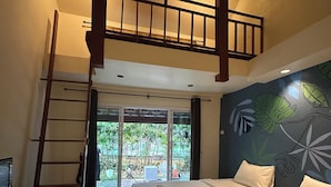 Ein Schlafzimmer mit Bett, Schreibtisch, einer Leiter und einem Balkon mit Blick auf Grünflächen.