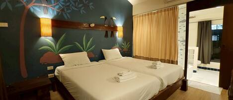 Habitación de hotel con dos camas, un mural de un árbol y pájaros, y vista al baño por una puerta abierta.