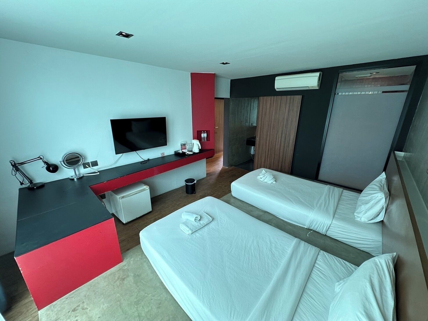 A hotel room with two beds, a desk with a flat-screen TV, a red accent wall, and a minibar.