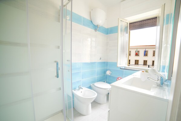 Baño con mampara de vidrio, bidé, inodoro y lavamanos con ventana arriba.