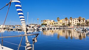 A marina with boats docked, a sailboat with a blue and white sail, and a row of buildings in the background. - FollowTheSun- Sea View House (Portoscuso)