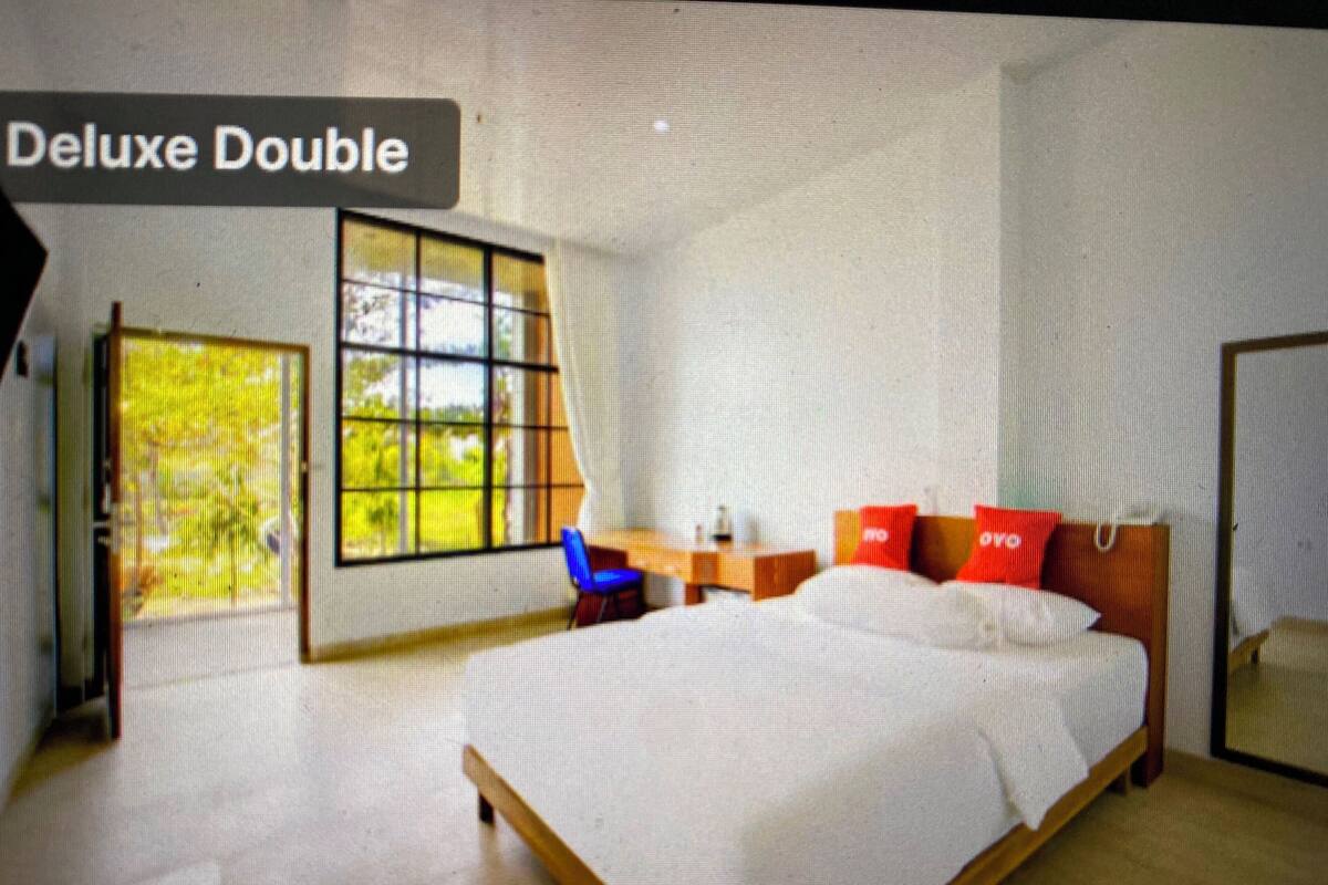 A room with a large bed, a desk, and a chair. There is a window with a view of greenery outside.