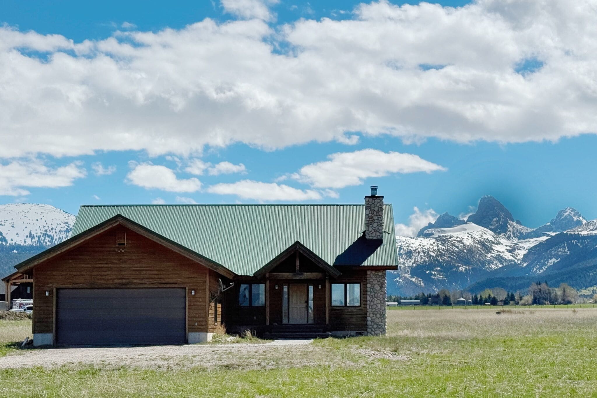 Une cabane en bois avec un toit vert et une cheminée en pierre, se dressant sur un fond de montagnes enneigées et d’un ciel dégagé.