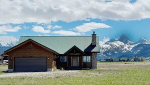 A wooden cabin with a green roof and stone chimney, set against a backdrop of snow-capped mountains and a clear sky.