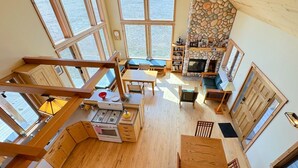 A spacious loft-style room with large windows, a stone fireplace, wooden furniture, and a kitchen area.