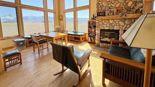 A spacious living area with large windows, a stone fireplace, wooden furniture, and a bookshelf.