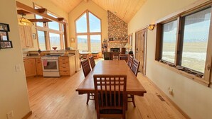 A spacious dining area with a long wooden table and chairs, a kitchen with wooden cabinets and a stone accent wall, and large windows offering a view of the outdoors.