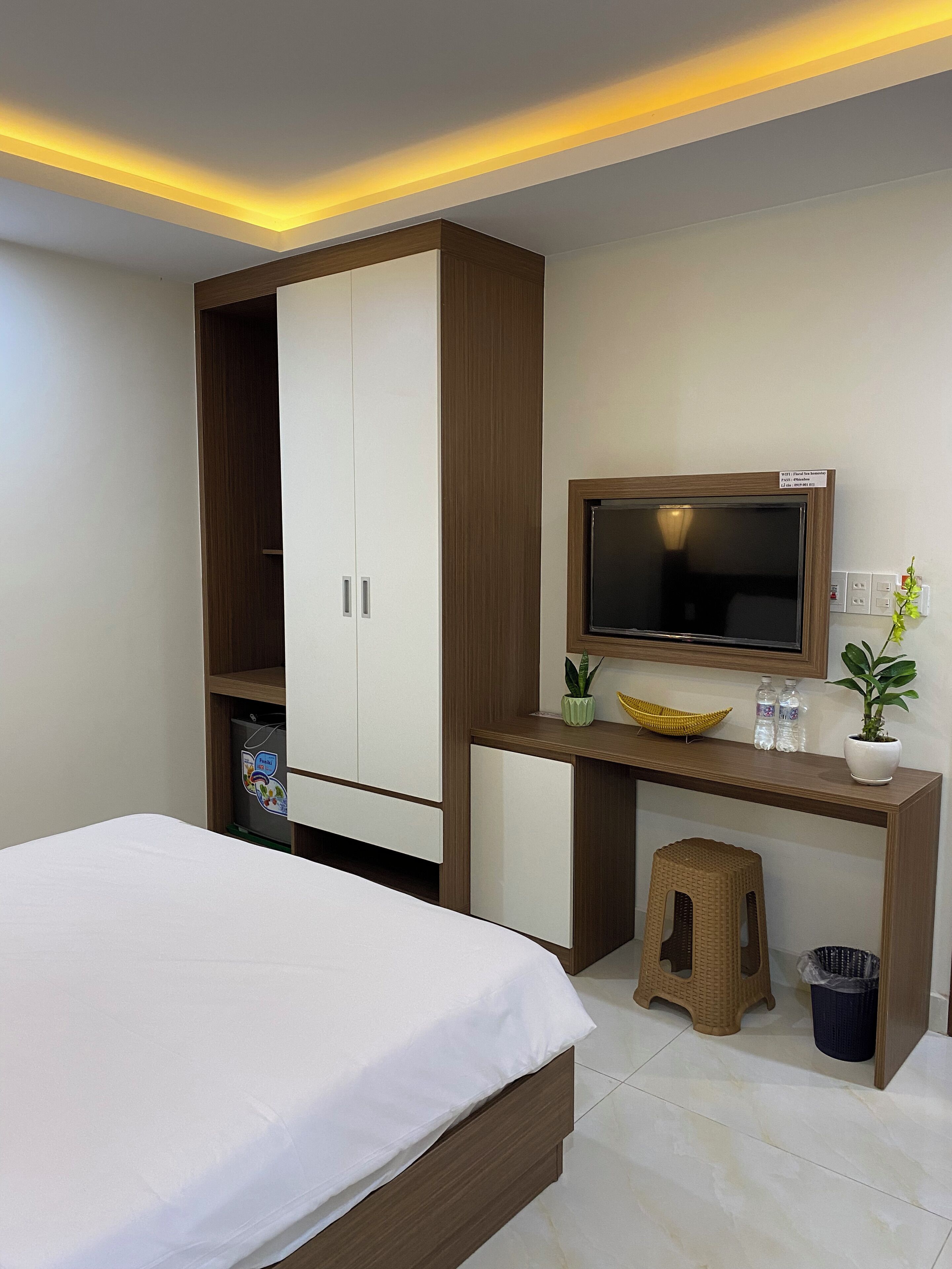A bedroom with a bed, a wardrobe, a desk with a TV, a small stool, and a trash bin.