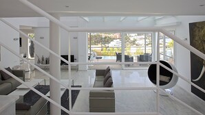 A modern living room with a view of a cityscape through large windows, a round seating area, and a staircase with white railings. - My Rental Homes - Villa Bridget with private pool and terraces (San Jose)