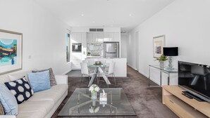 A modern living room with a glass coffee table, a sofa with patterned cushions, a TV on a wooden stand, and a dining area with a glass table and chairs. - Swainson on Hindmarsh (Adelaide)
