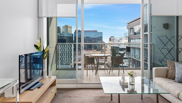 A modern living room with a large TV, glass coffee table, and a balcony with a city view. - Swainson on Hindmarsh (Adelaide)